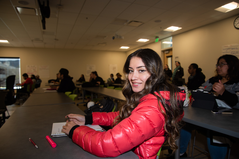 Student wearing a red jacket seating in a classroom smiles while looking at the camera