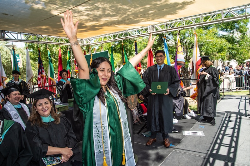 A graduate in regalia raises her hands in celebration on stage, surrounded by smiling staff.