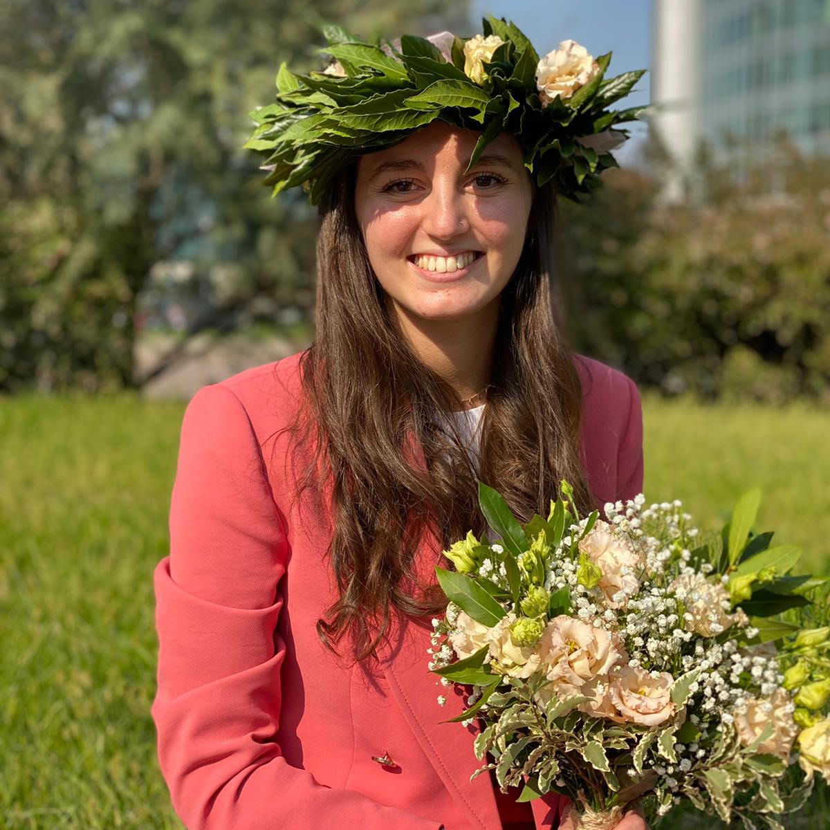 woman with long chestnut brown hair, smiling at the camera. She has flowers in her hair and hands. 