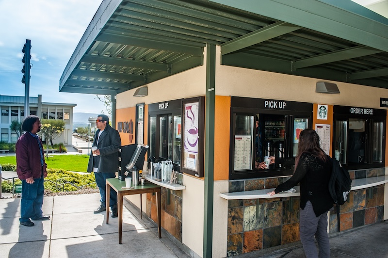 A Pony Espresso barista hands a coffee in a paper cup to a student. Nearby, two students stand outside the building, chatting and sipping their beverages.