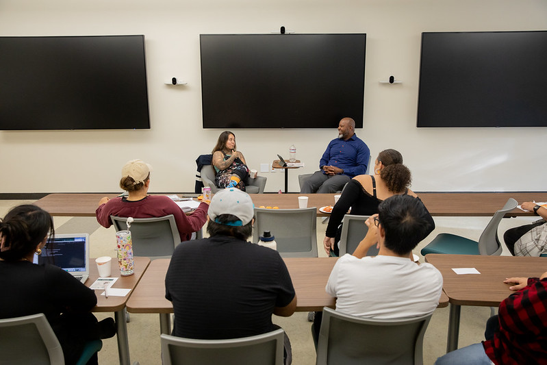 A presenter speaks to an audience of students seated at tables during an event.
