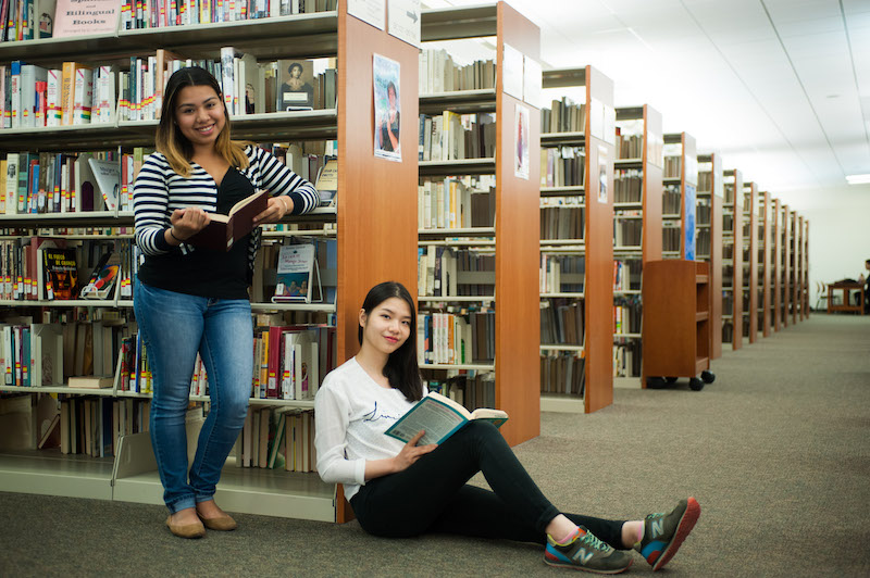 Two students in the college library, one standing and the other seated on the floor, both with open books, smiling, and looking at the camera. One student is leaning against a bookshelf.